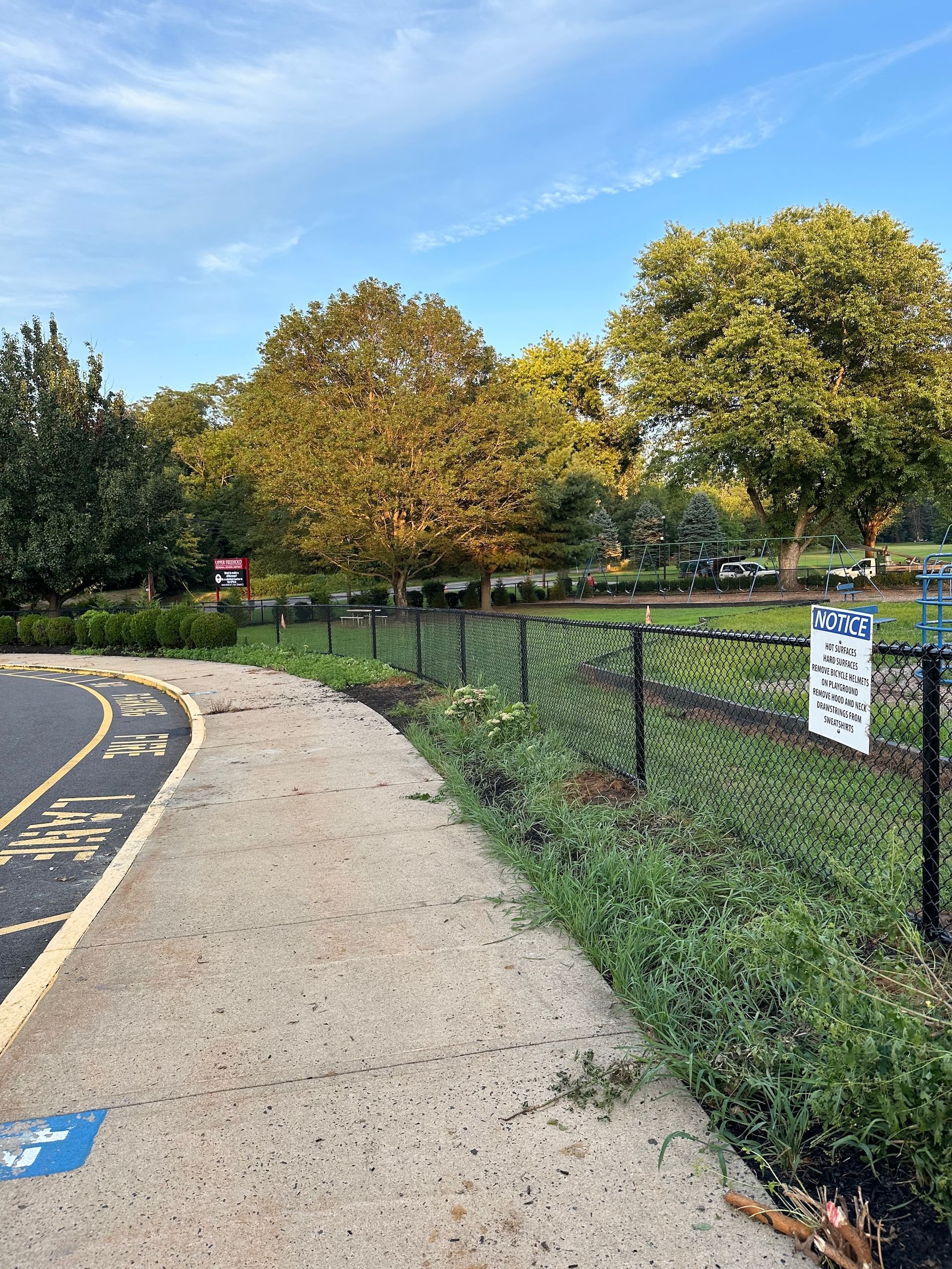 Pathway curves along a fenced-in park with trees and a sign, under a blue sky.