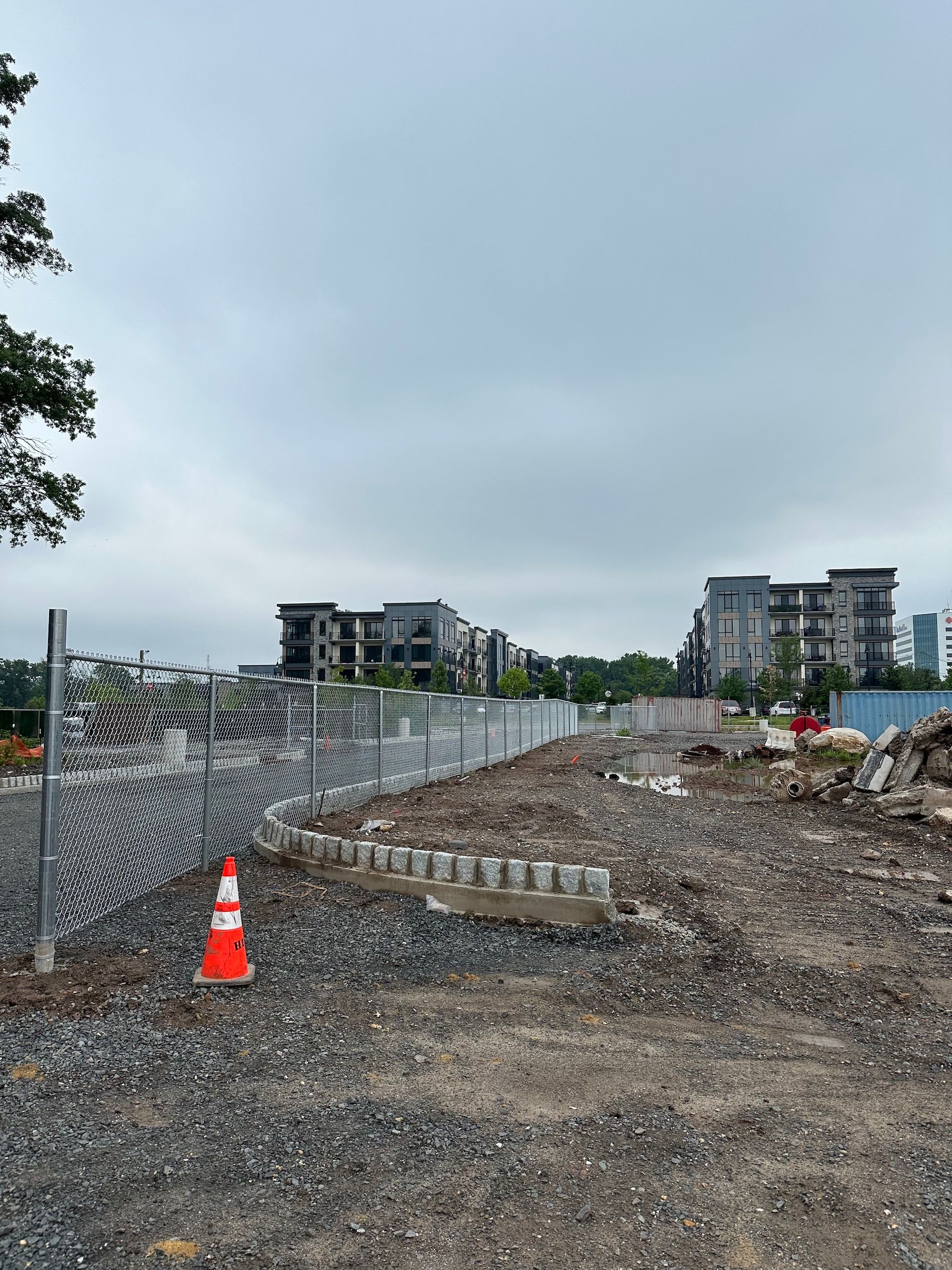 Construction site with a chain-link fence, gravel, concrete blocks, and buildings in the background under a cloudy sky.