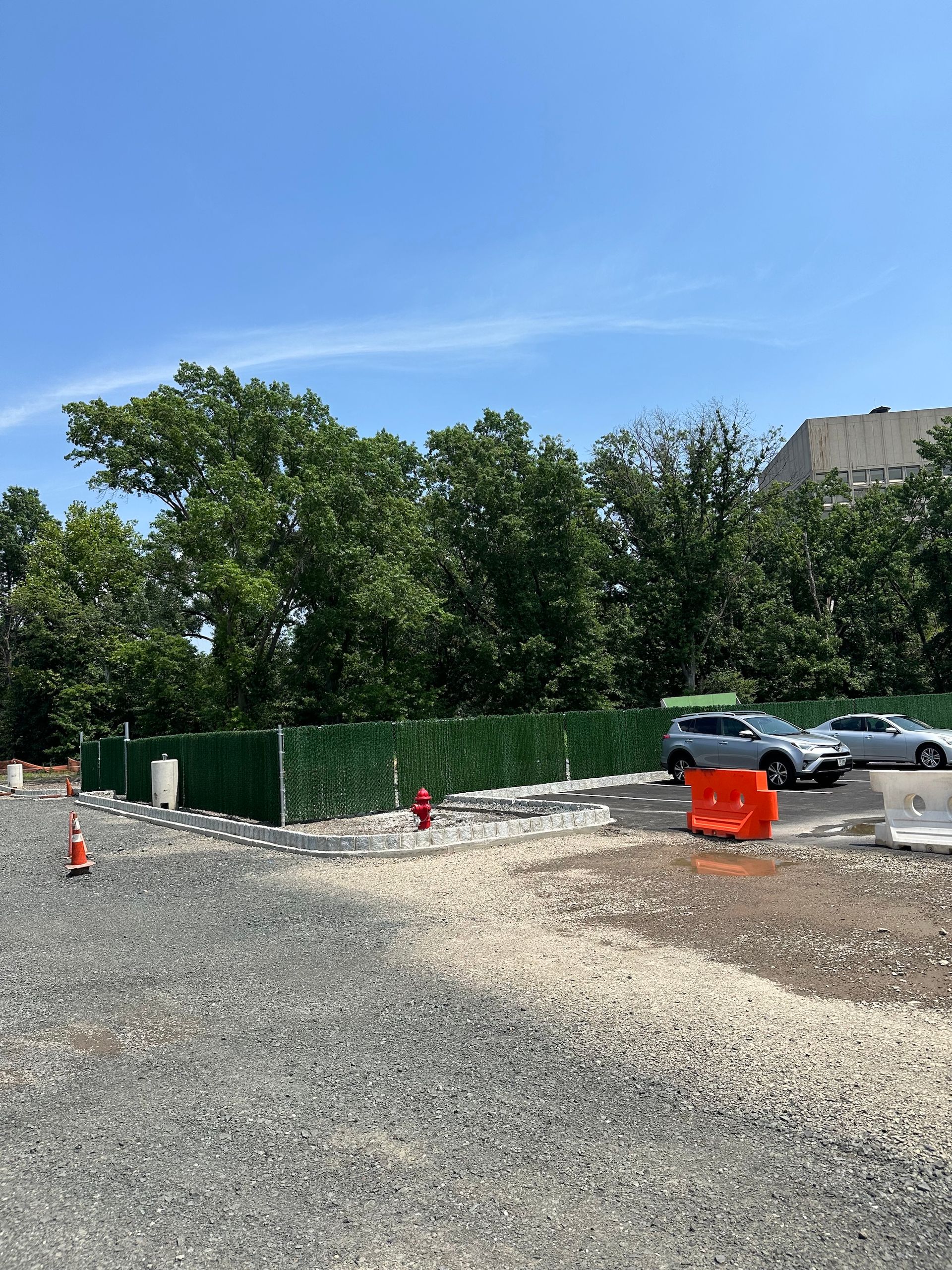 Construction site with gravel, a green fence, trees, cars, and a blue sky.