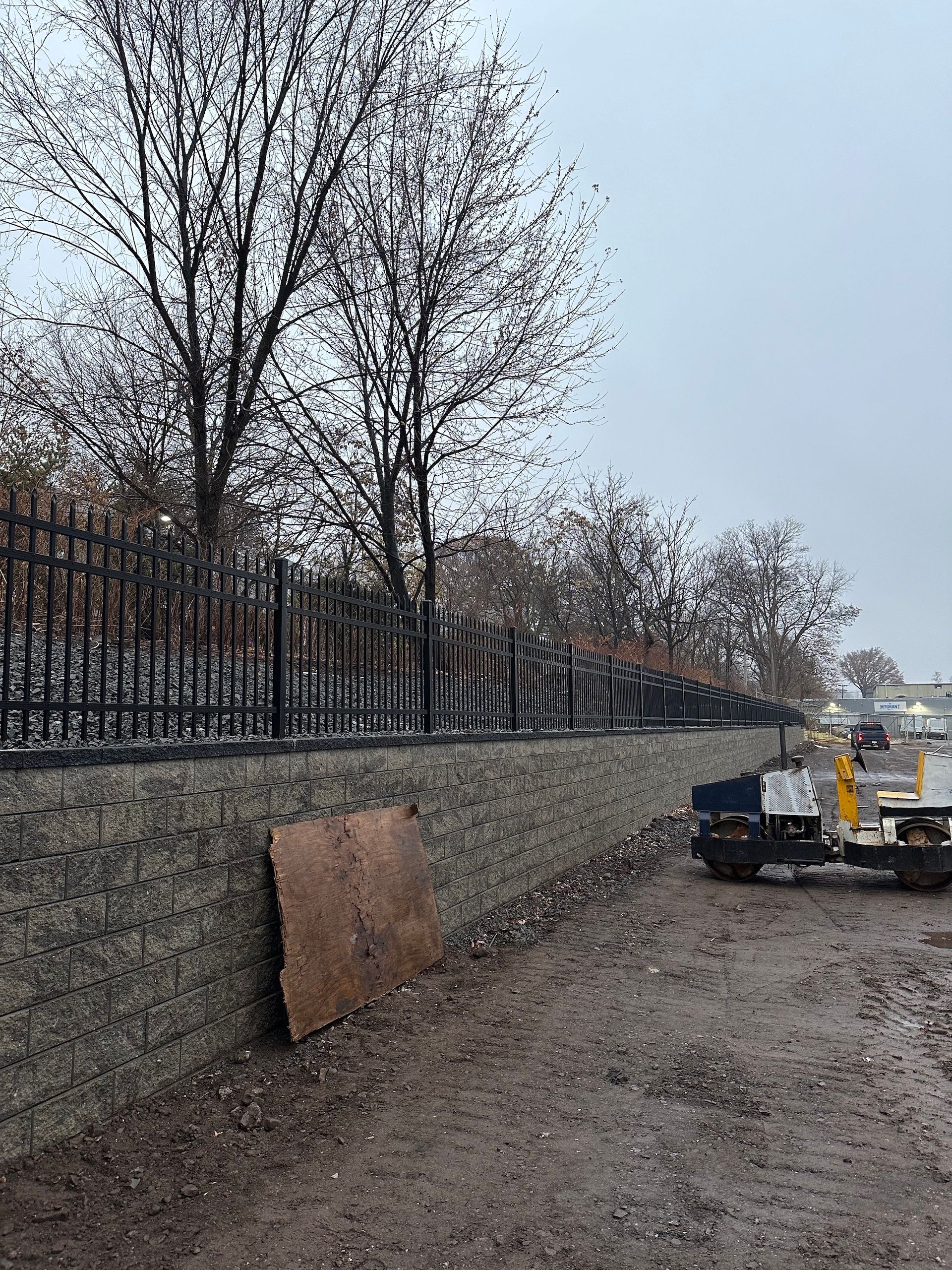 Black fence atop a retaining wall, bare trees, overcast sky, construction equipment on muddy ground.