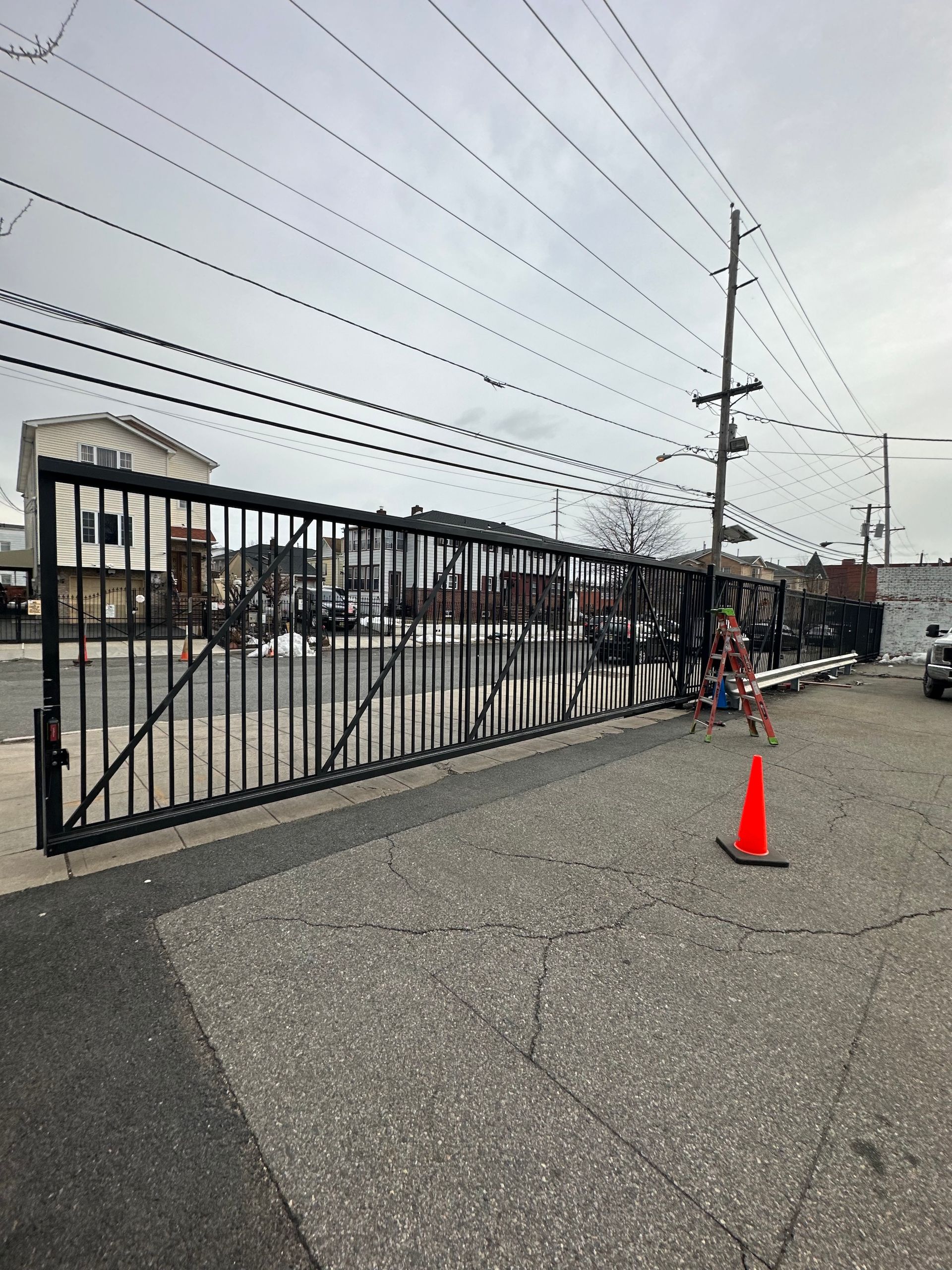 Black metal gate on asphalt, orange cone, power lines, and building. Gray sky in background.