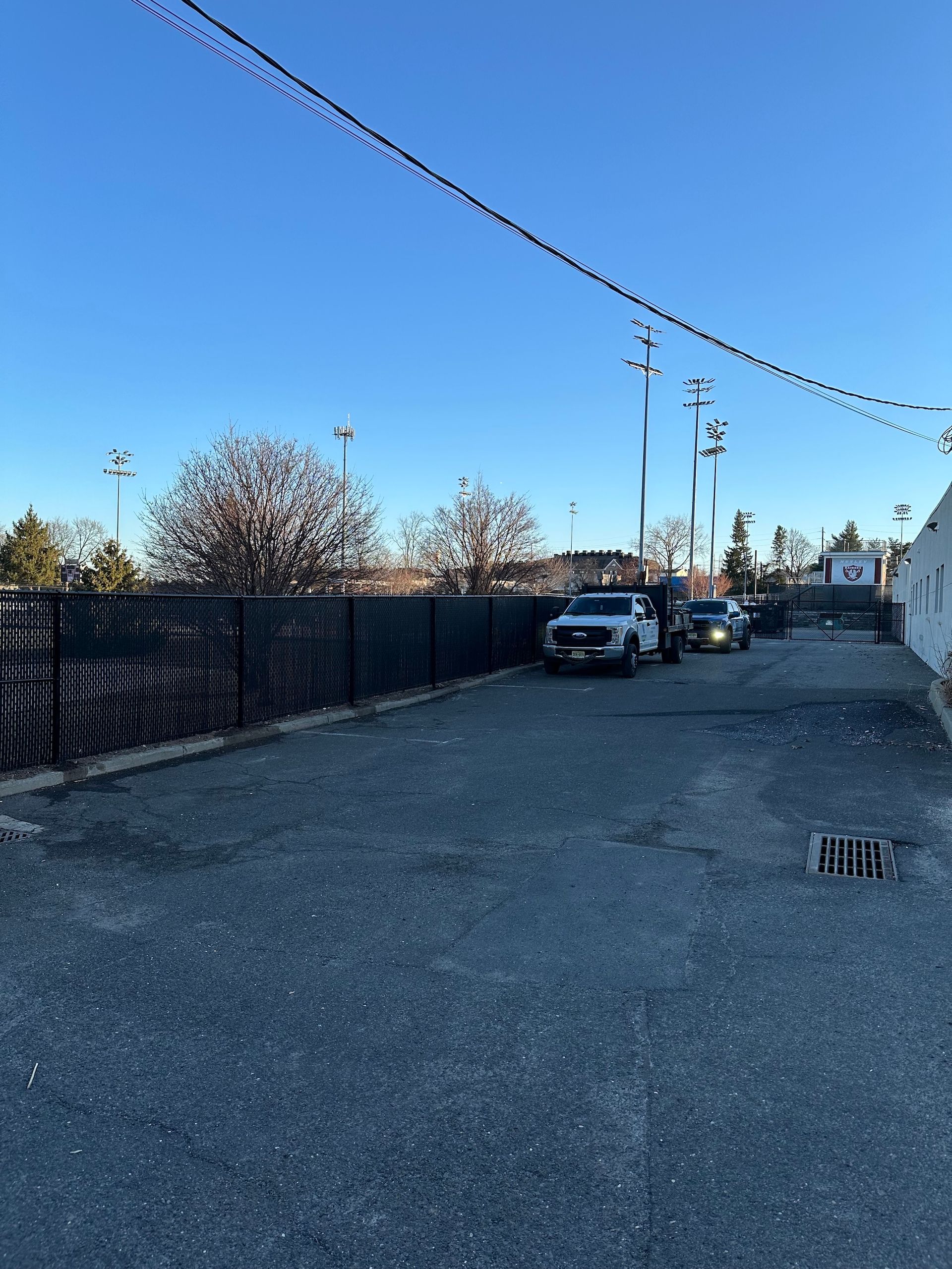 Gravel alley with vehicles. A black fence lines the left. Power lines and a flowering tree are visible in the background.