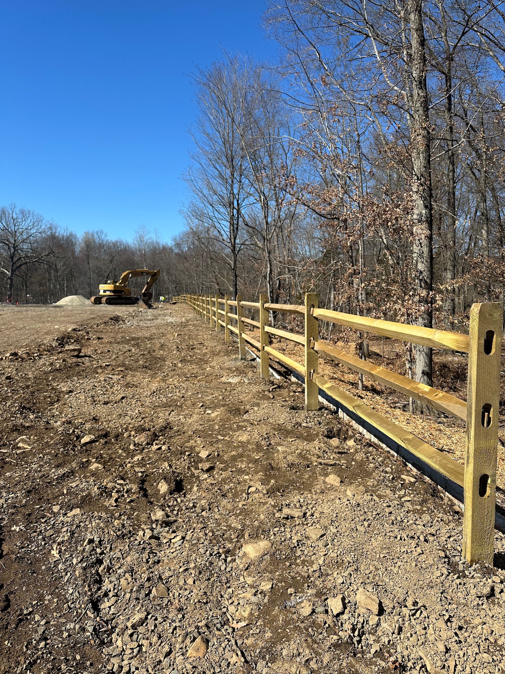 Wooden split-rail fence alongside a dirt path, with an excavator in the background and trees under a blue sky.