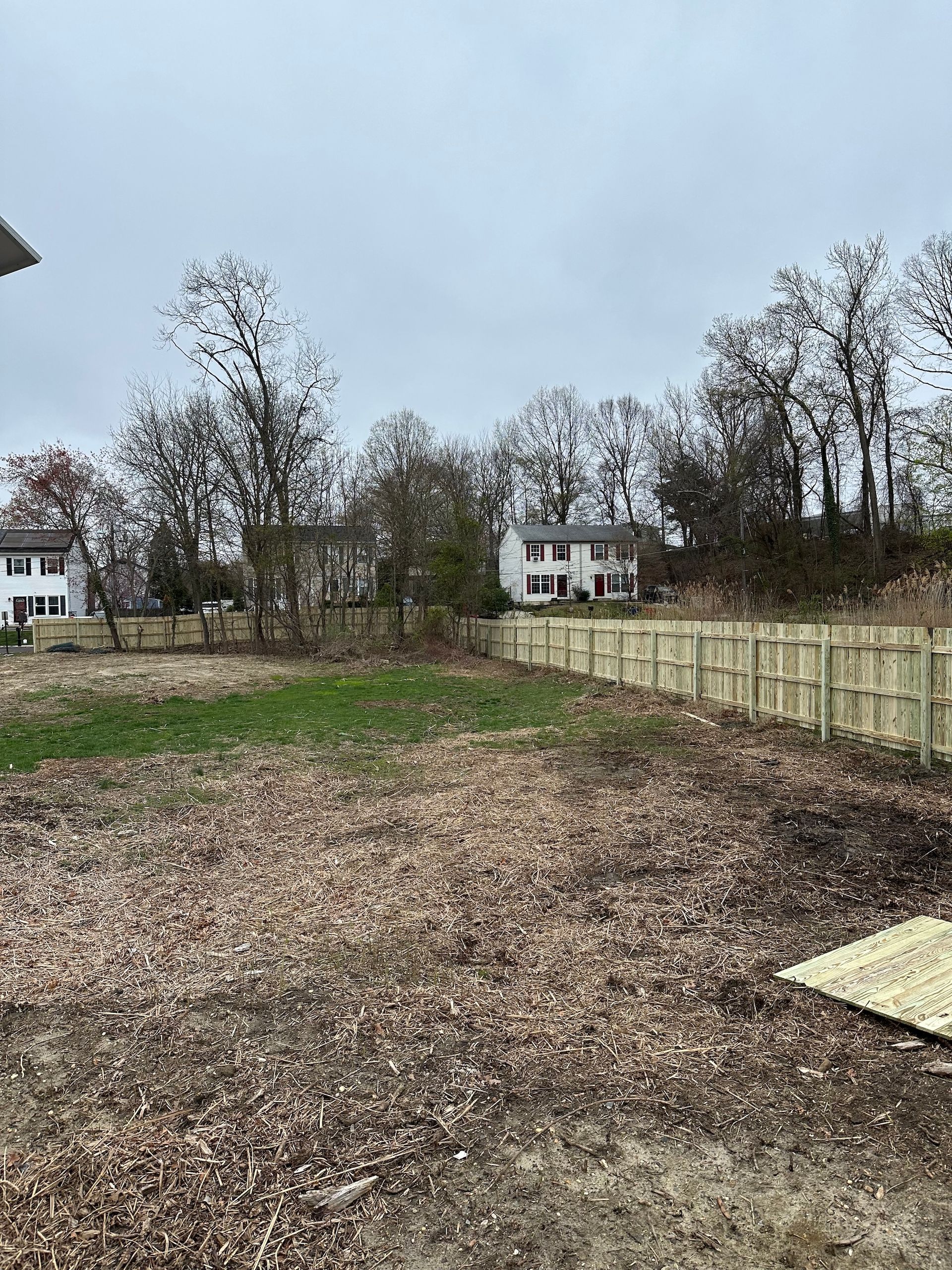 A vacant lot covered in fallen leaves, with a new wooden fence, trees, and houses in the background on a cloudy day.