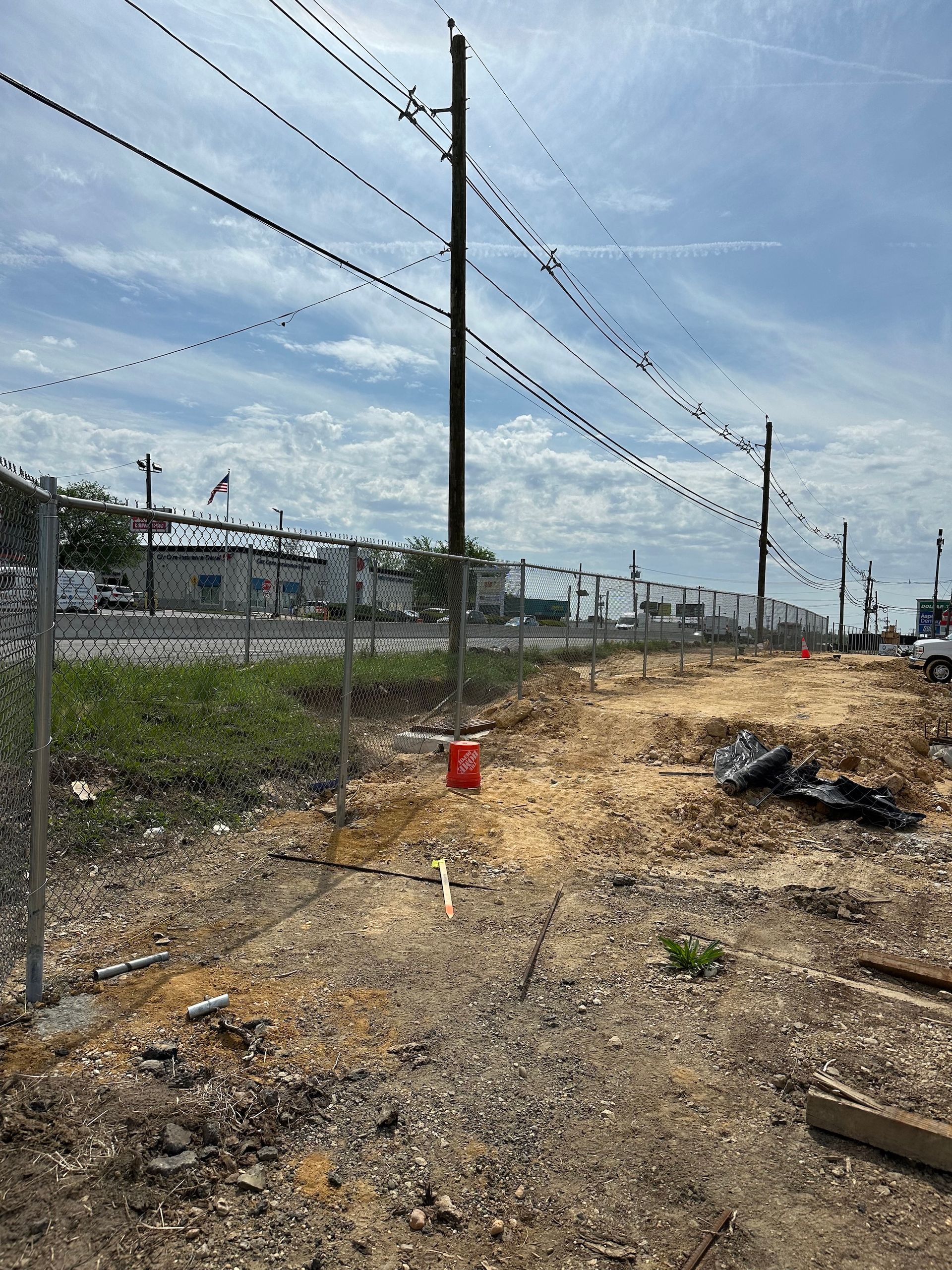 Chain-link fence borders dirt path, power lines overhead, industrial buildings in the background, blue sky.