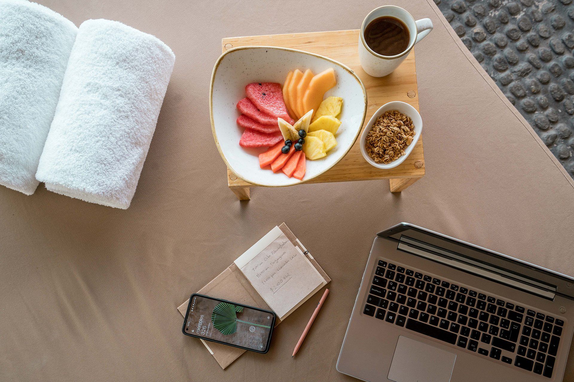 Hay una computadora portátil sobre una cama al lado de un bol de fruta y una taza de café.