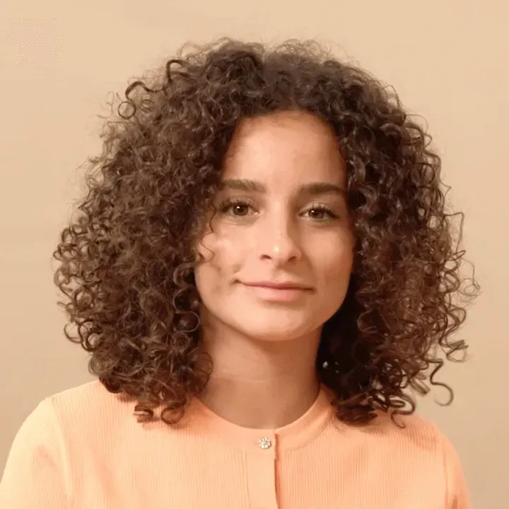 Una mujer con cabello rizado lleva una camisa rosa y sonríe.