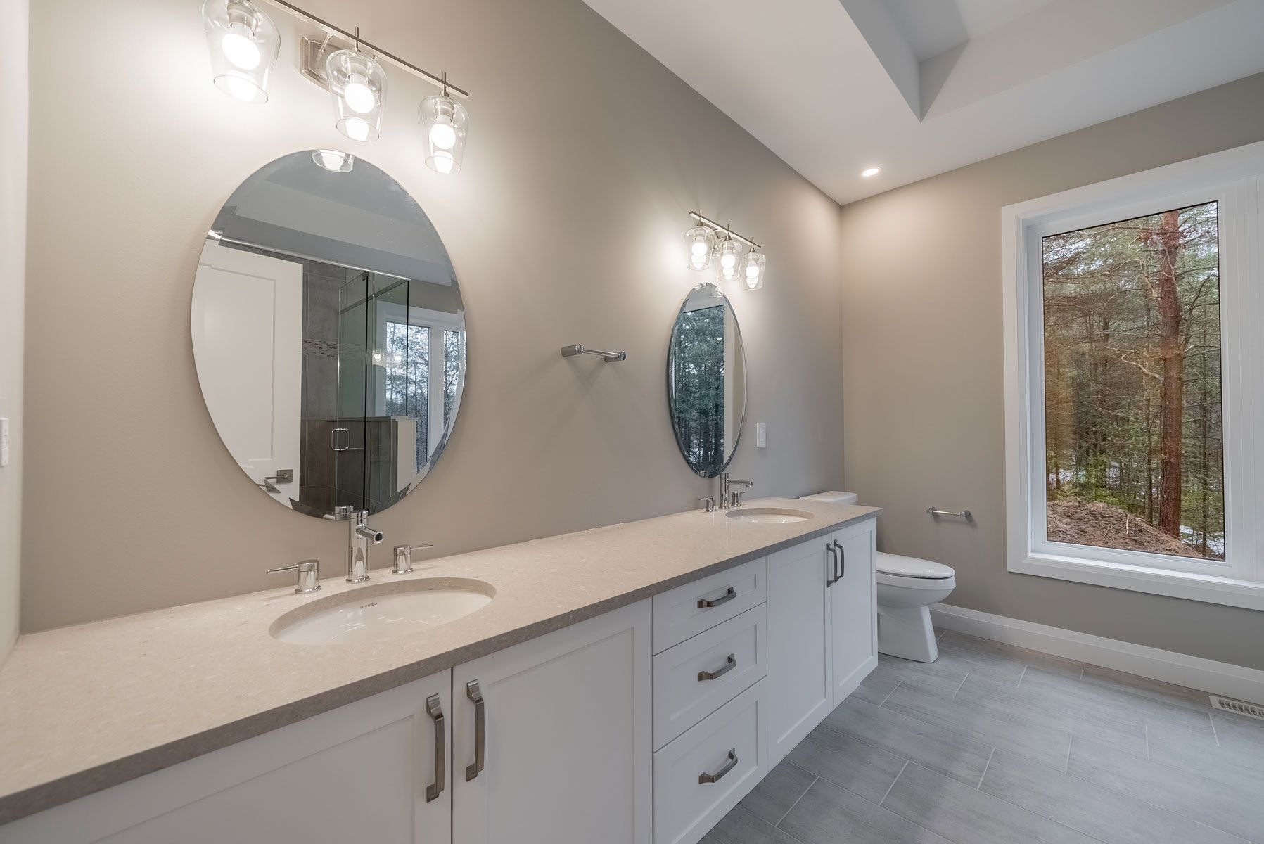 A bathroom with two sinks , two mirrors , a toilet and a window  in Fidelity Homes Signature build in Cramahe Township