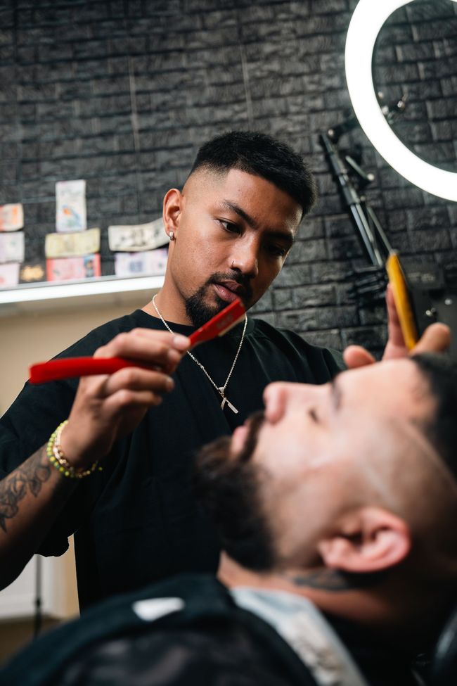 Barber trimming a client’s beard in a dim salon, with a ring light and brick wall behind them