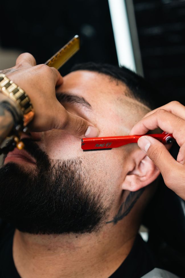 Barber trimming a man's beard with a red razor close to his ear