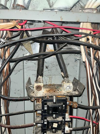 Open electrical breaker panel with tangled black, red, and beige wires and rusted components.
