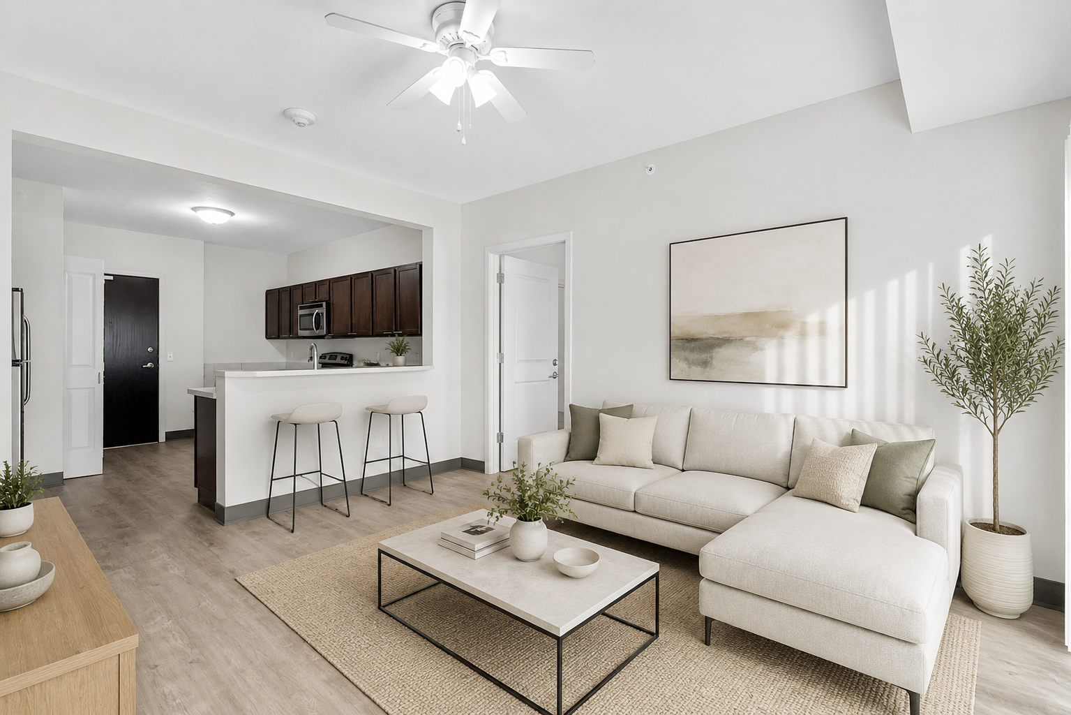 Living room with brown sofa, coffee table, and open kitchen with bar stools. Hardwood floors and white walls.