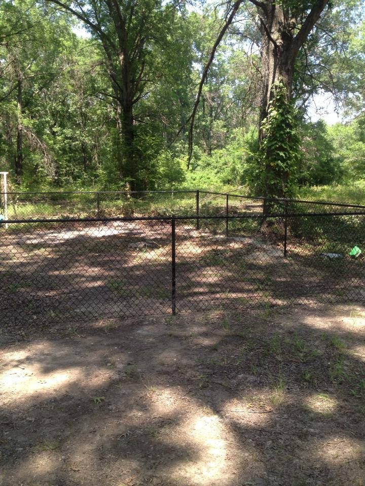 A chain link fence surrounds a dirt field with trees in the background.