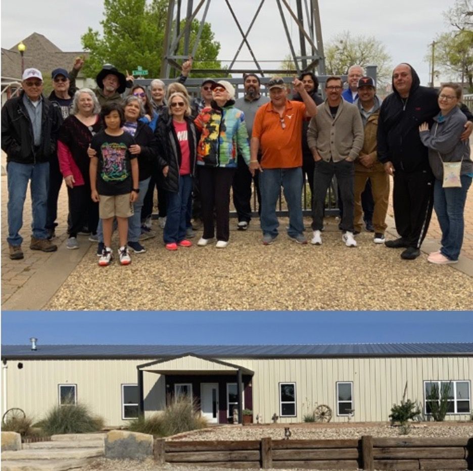 Group of people posing by a windmill, with a building below. They appear to be outdoors on a sunny day.