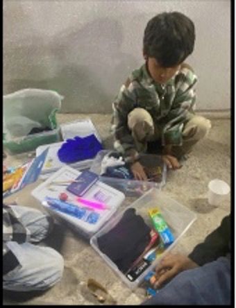 Boy in camouflage crouches, sorting through supplies in containers on the floor, indoors.