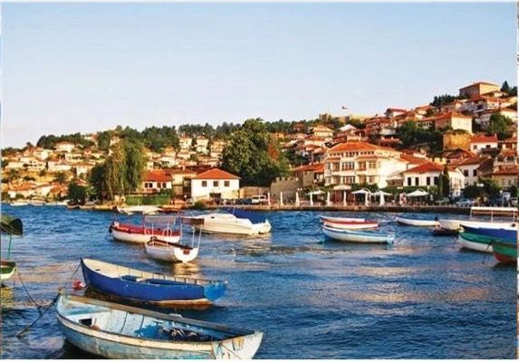 Boats bob in a harbor. Buildings with red-tiled roofs dot a hillside. Bright blue water.