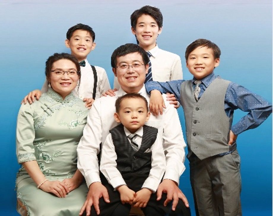 Family portrait: six people posing against blue backdrop. Smiling, formal attire.