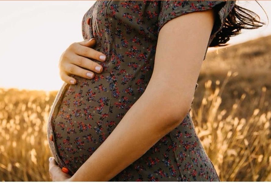 Pregnant person in a floral dress, hands cradling belly, standing in a field with warm sunlight.
