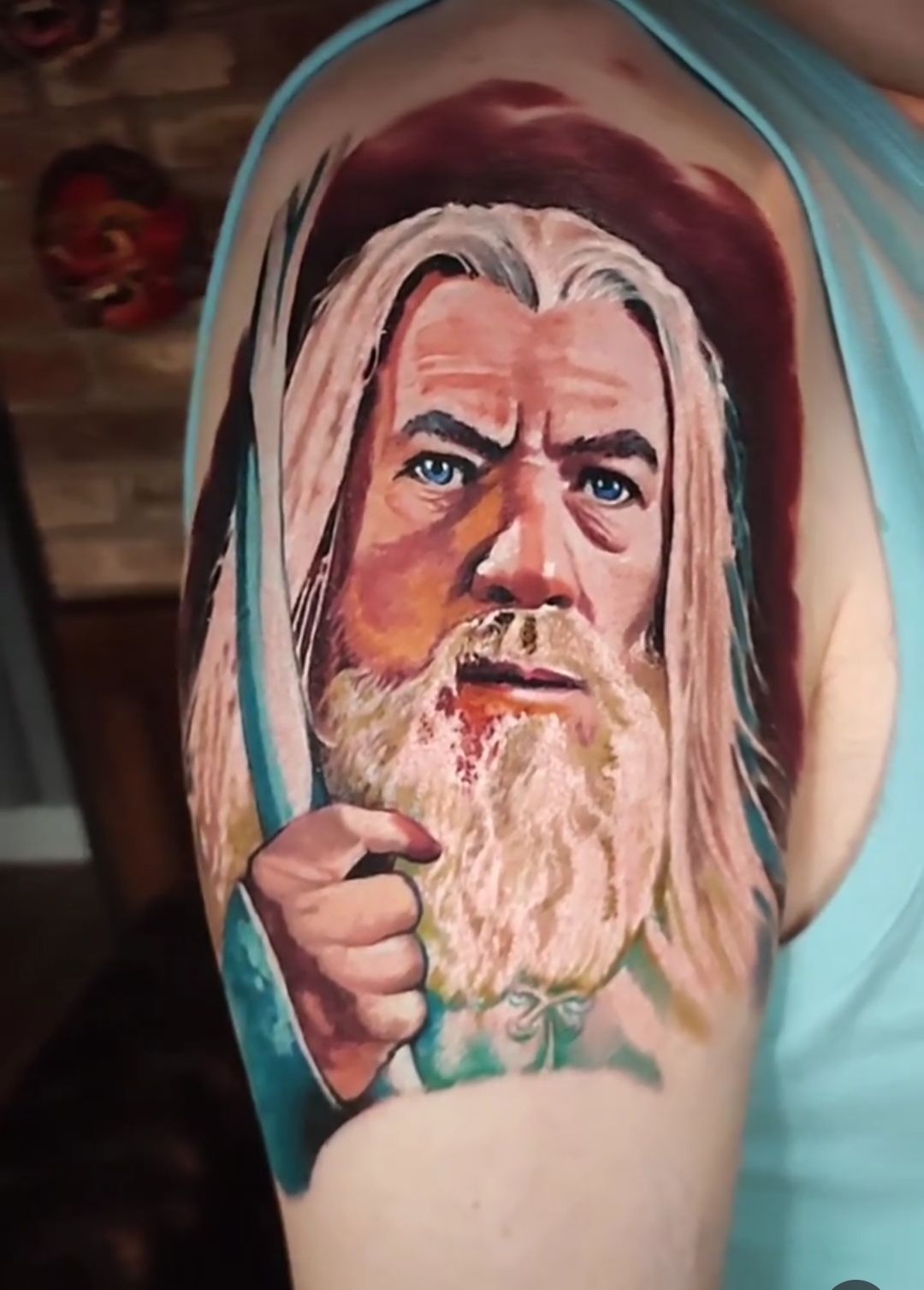Gandalf tattoo on upper arm, holding staff. White beard, blue eyes, red background.