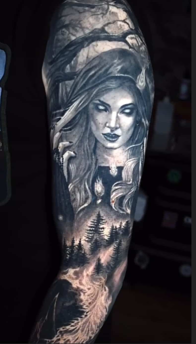 Tattoo of a woman with white eyes, trees, and forest on an arm in black and gray.