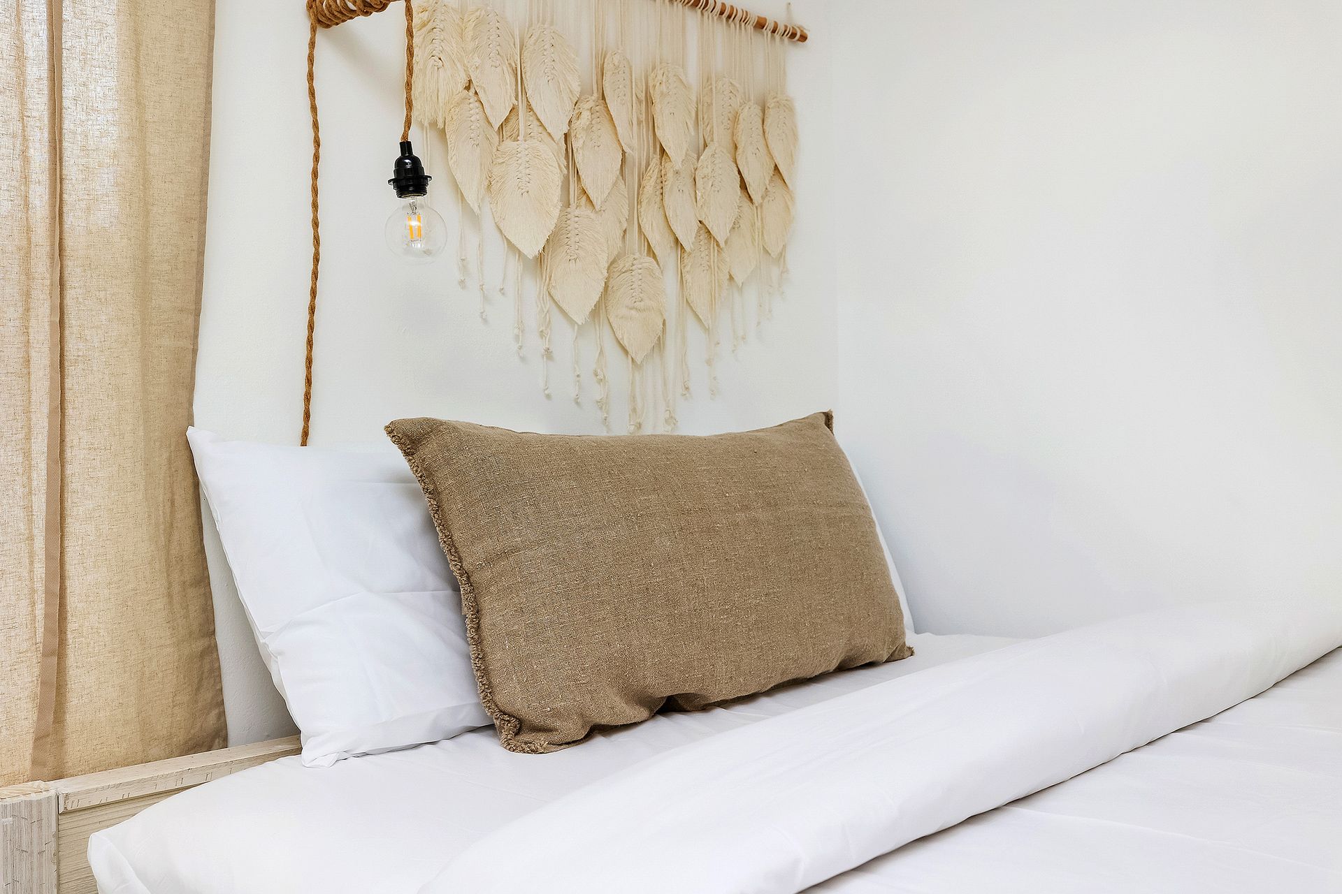 Bed with white linens, tan pillow, macrame wall hanging, and light fixture.