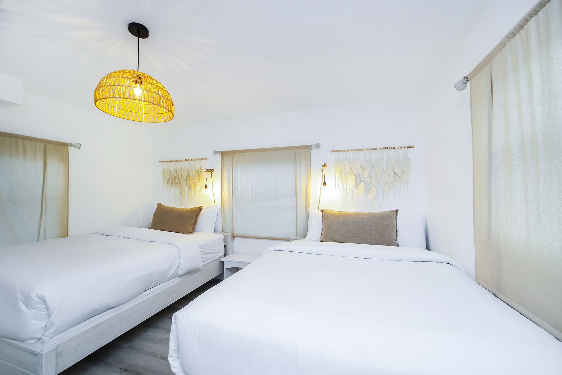 A bright white bedroom with two beds, woven lamp, neutral curtains, and decorative wall hangings.