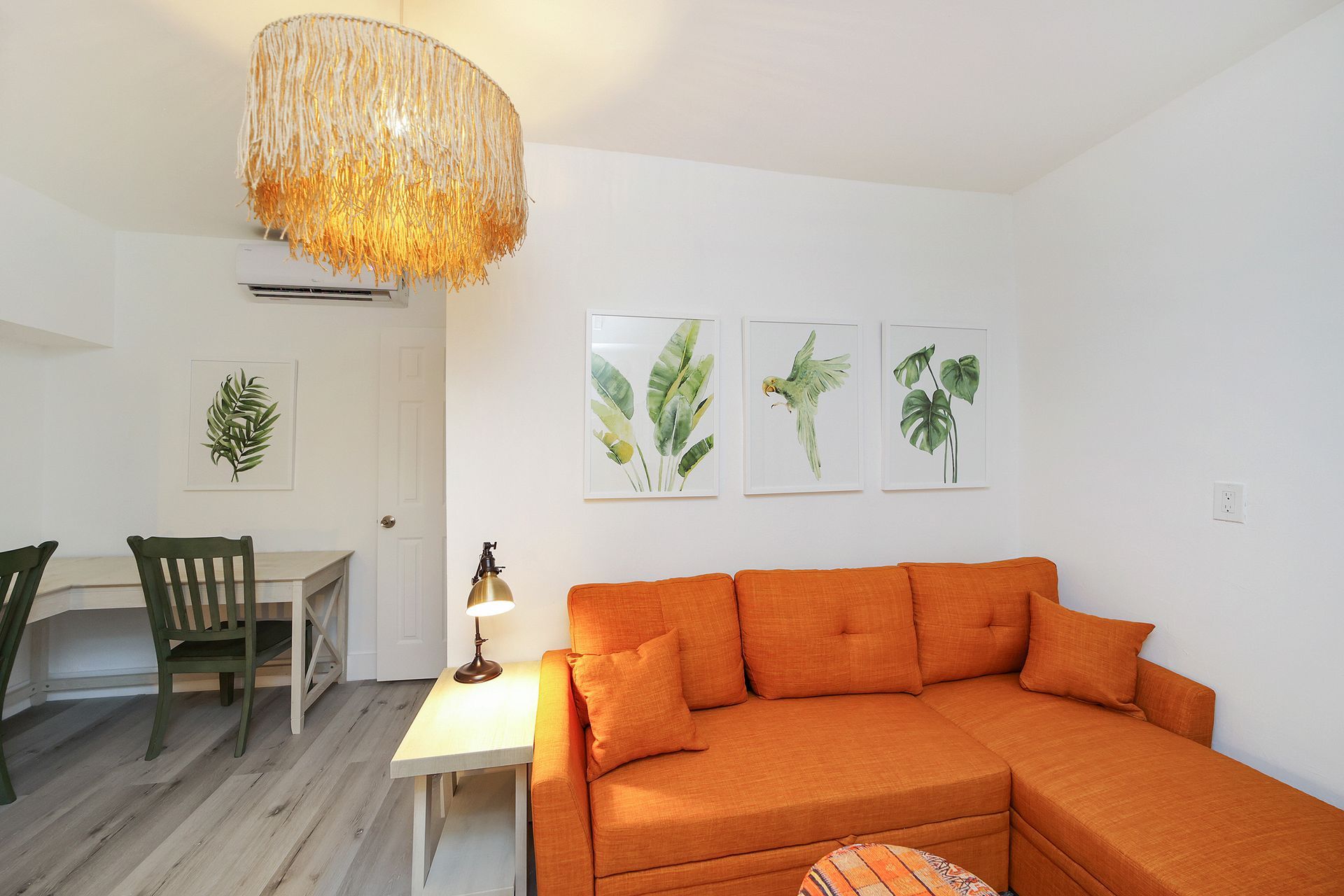 Orange couch in a brightly lit room with botanical art, a desk, and a unique chandelier.