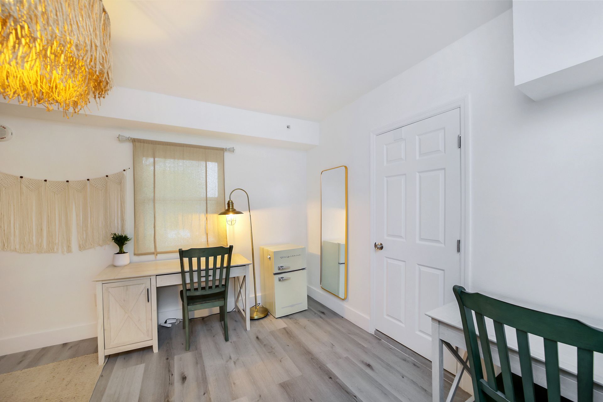 A room with a desk, chair, mirror, and a white door; a gold chandelier hangs from the ceiling.