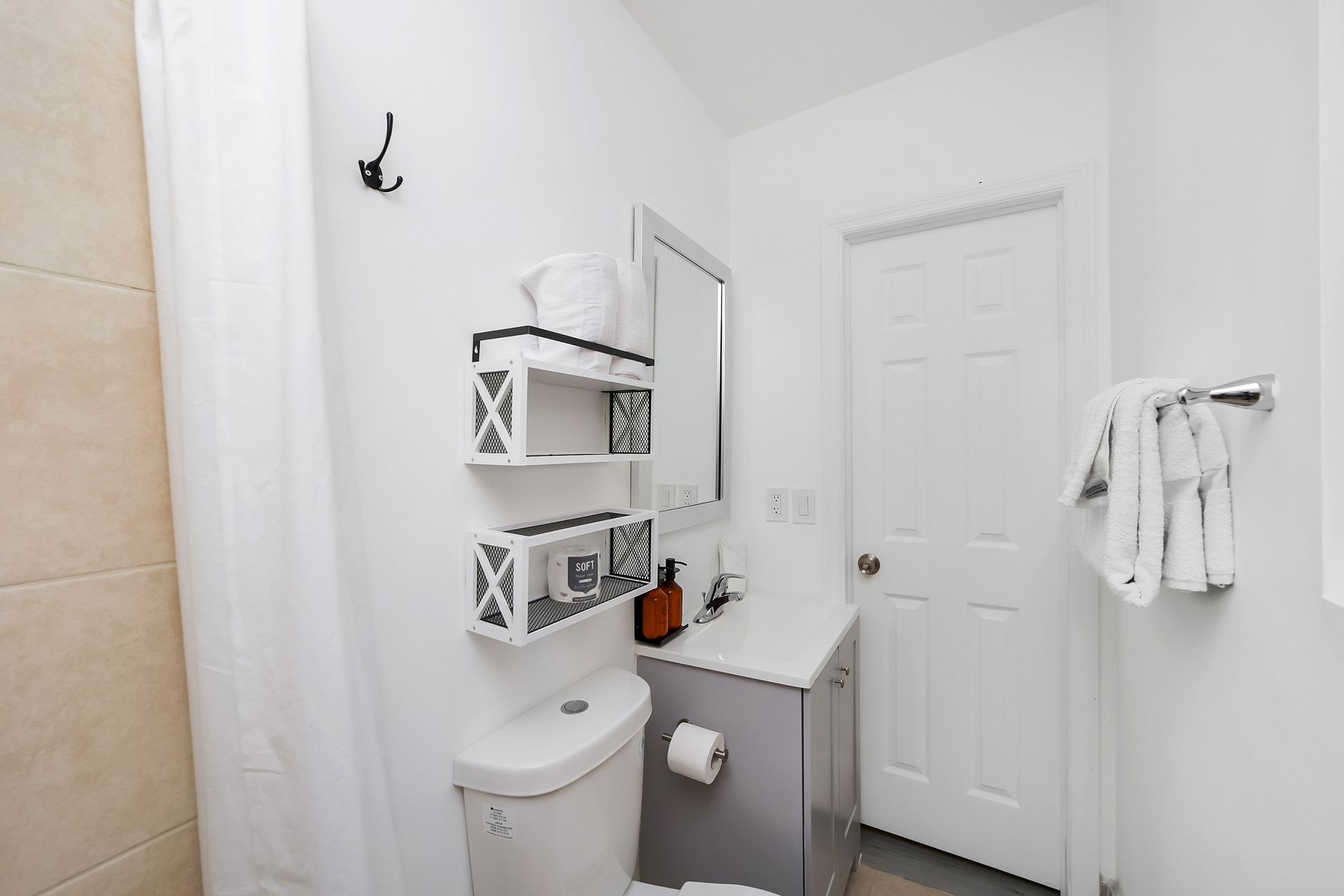 White bathroom with toilet, sink, mirror, and shelves. White door on the right. Beige shower tiles.
