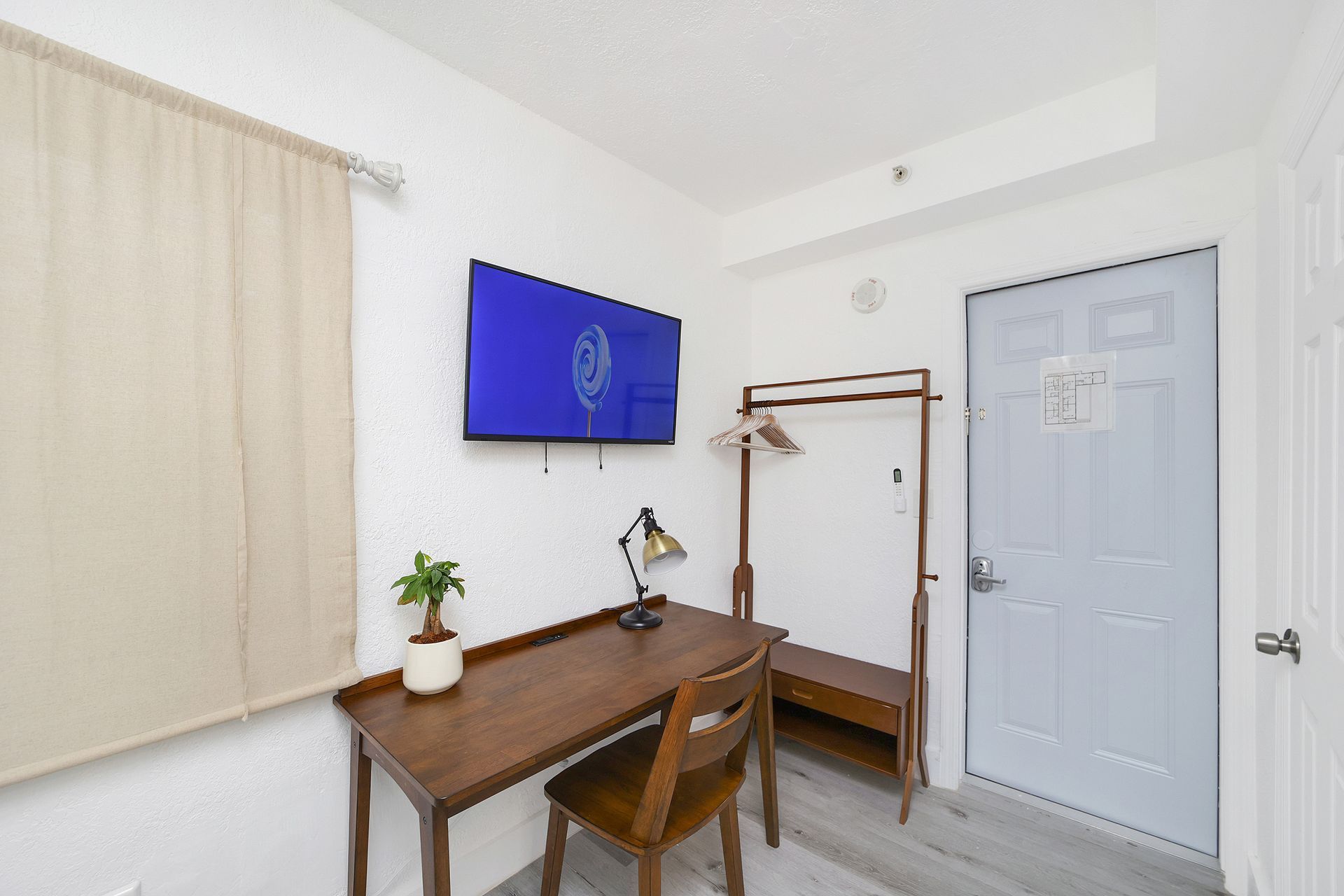 Hotel room with desk, TV, open closet, and closed door. Off-white walls, gray floors, and beige curtains.