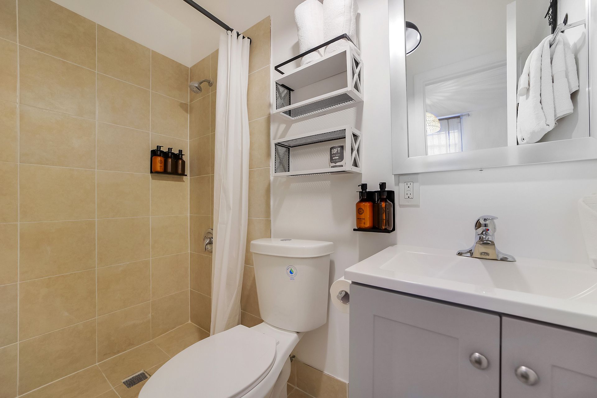 Small bathroom with beige tile, white toilet, vanity, and shower. Shelving unit holds toiletries.