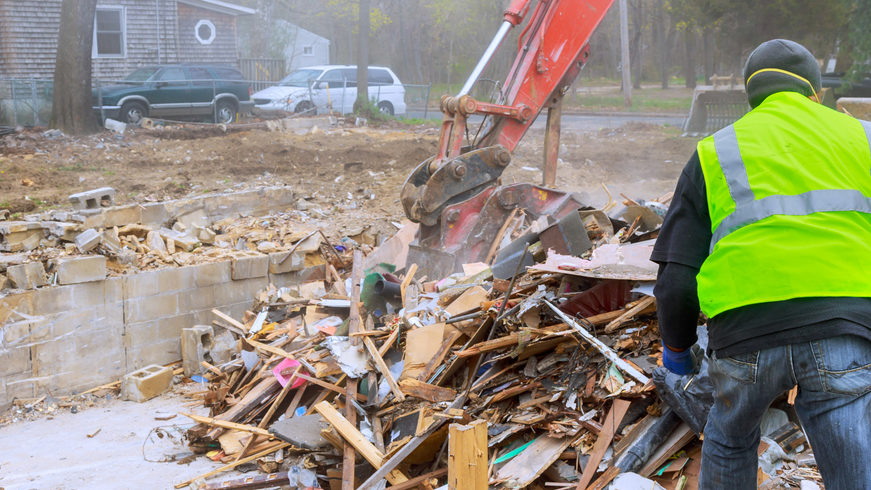Worker in a neon vest stands by an excavator clearing rubble at a demolition site.