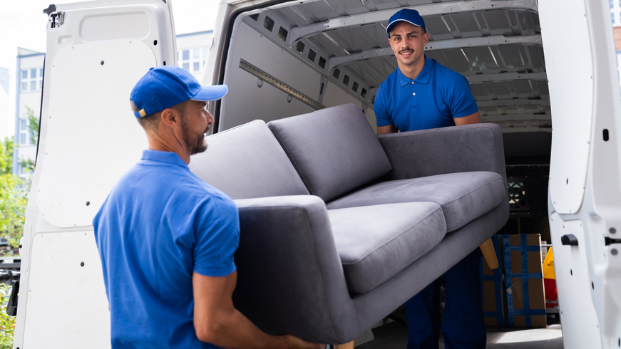 Two movers in blue uniforms carrying a gray couch into a white moving truck.