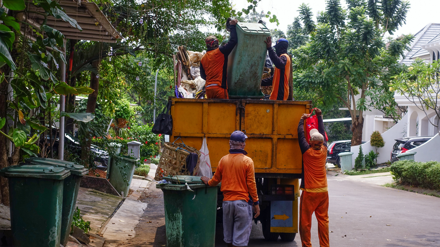 Workers emptying garbage bins beside a yellow trash truck on a leafy residential street