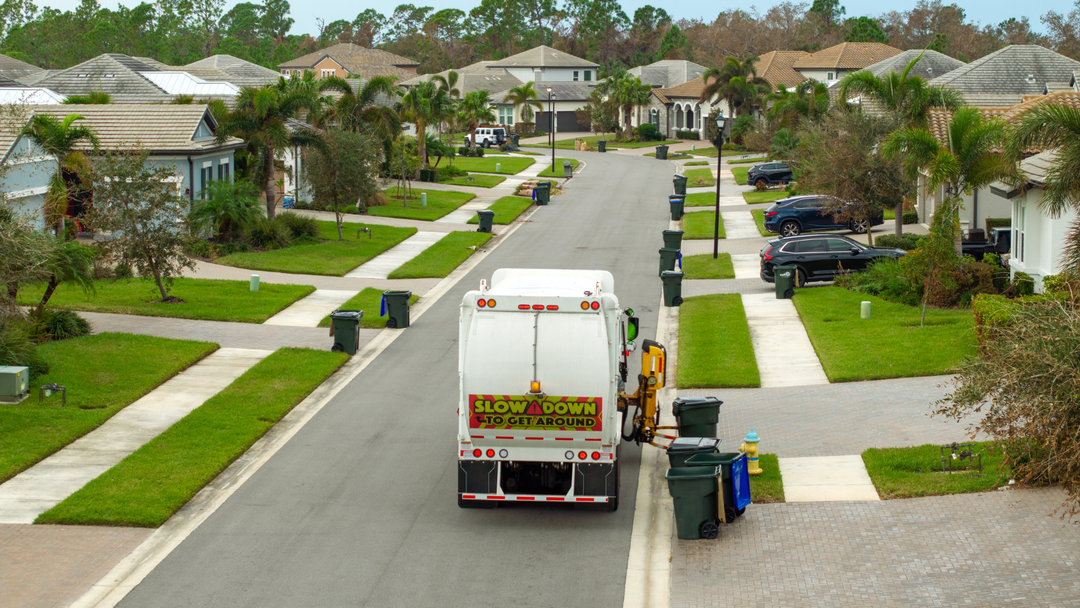 Garbage truck collecting bins on a suburban street with houses and lawns.