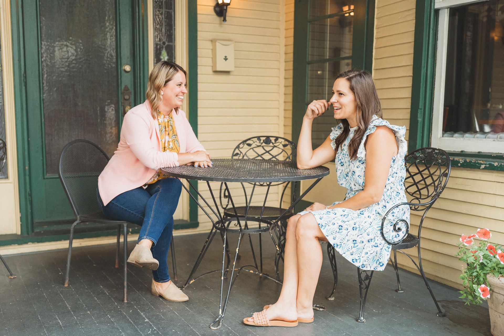 Two women are sitting at a table on a porch talking to each other.