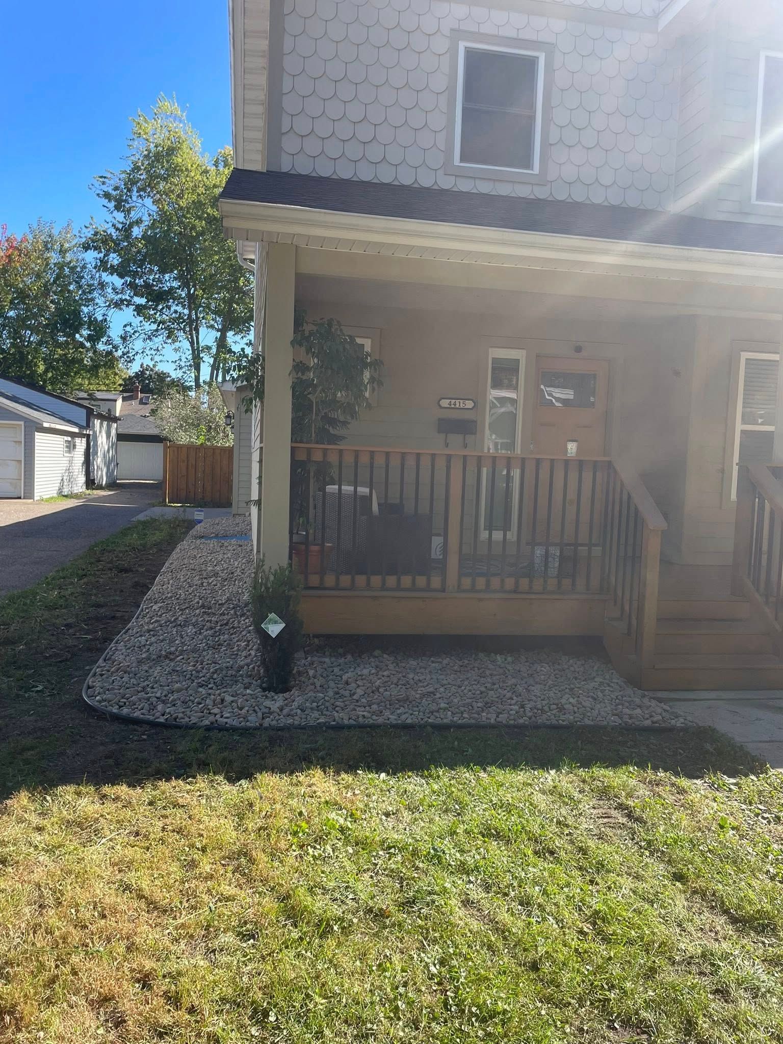 A beige two-story house with a wooden front porch and a gravel garden bed in the front yard under a clear blue sky.