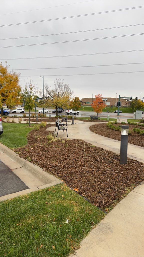 A paved walkway winds through a landscaped area with mulch and trees during an overcast autumn day.