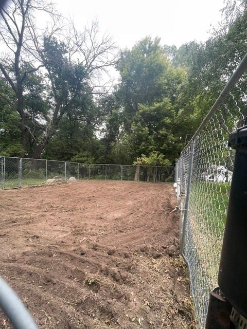 Freshly graded dirt lot beside chain-link fences and trees under an overcast sky