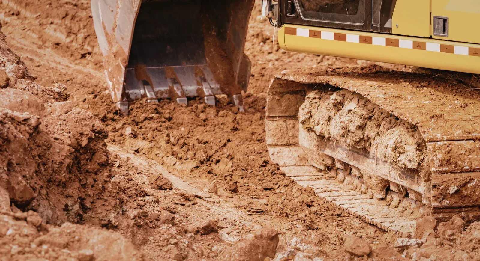 A close-up of a yellow excavator's metal bucket digging into brown soil next to its large, muddy tracked wheels.