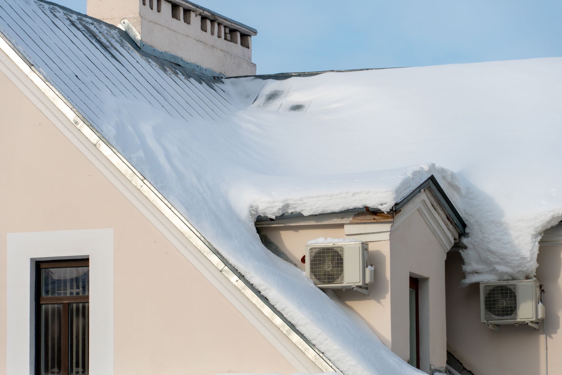 Snow-covered house roof with two wall-mounted air conditioners under a blue sky