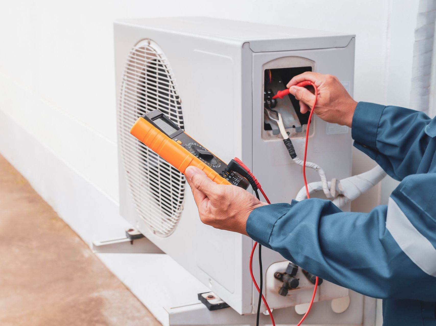HVAC technician in blue uniform, testing an outdoor air conditioning unit with a multimeter.