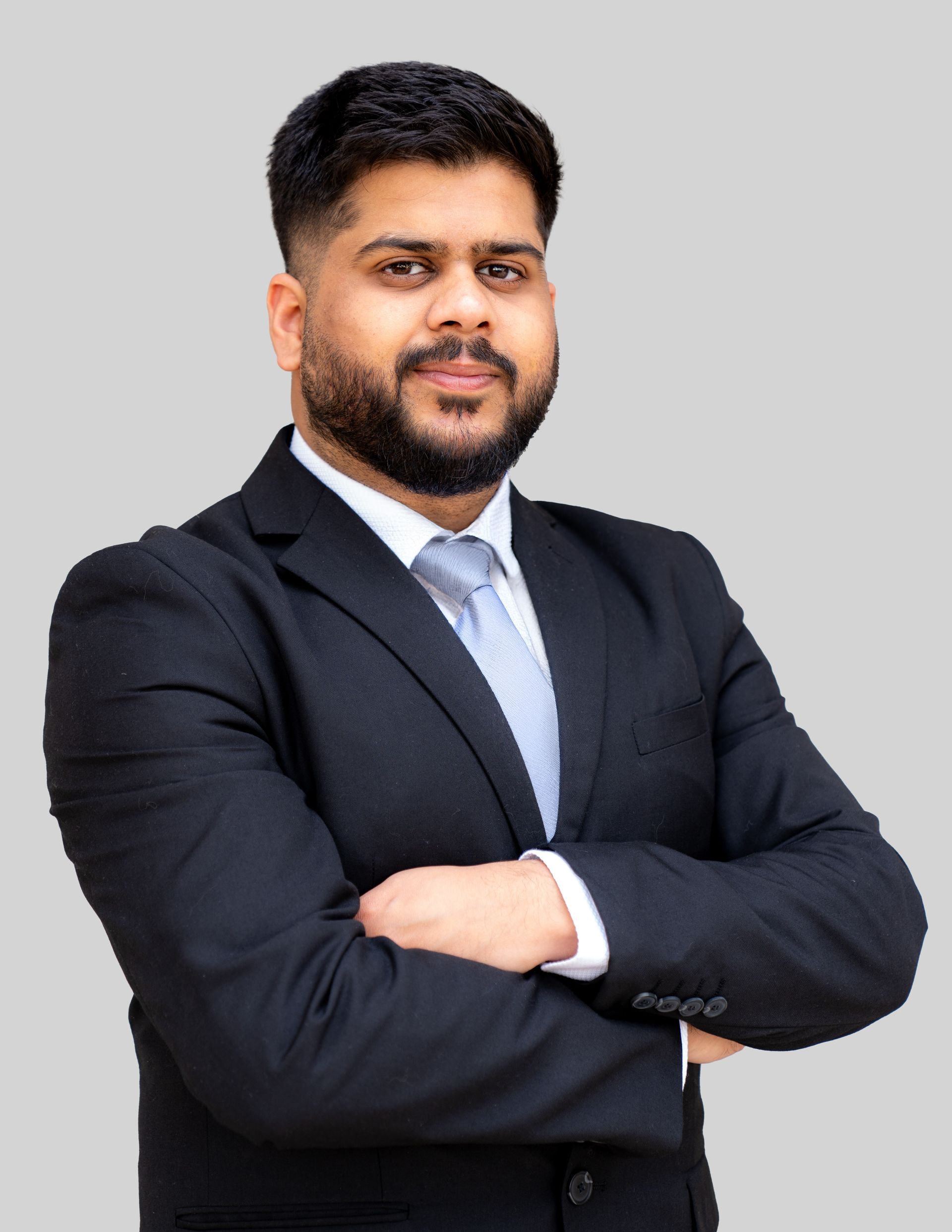 Professional portrait of a person in a black suit and light blue tie, standing with arms crossed against a gray background