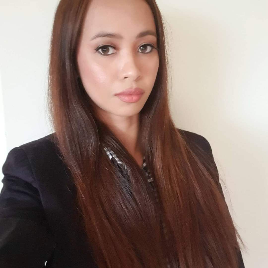 Woman with long straight brown hair wearing a black blazer, posing against a plain light wall.