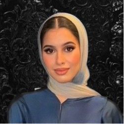 A woman wearing a hijab and a blue shirt is standing in front of a black background.