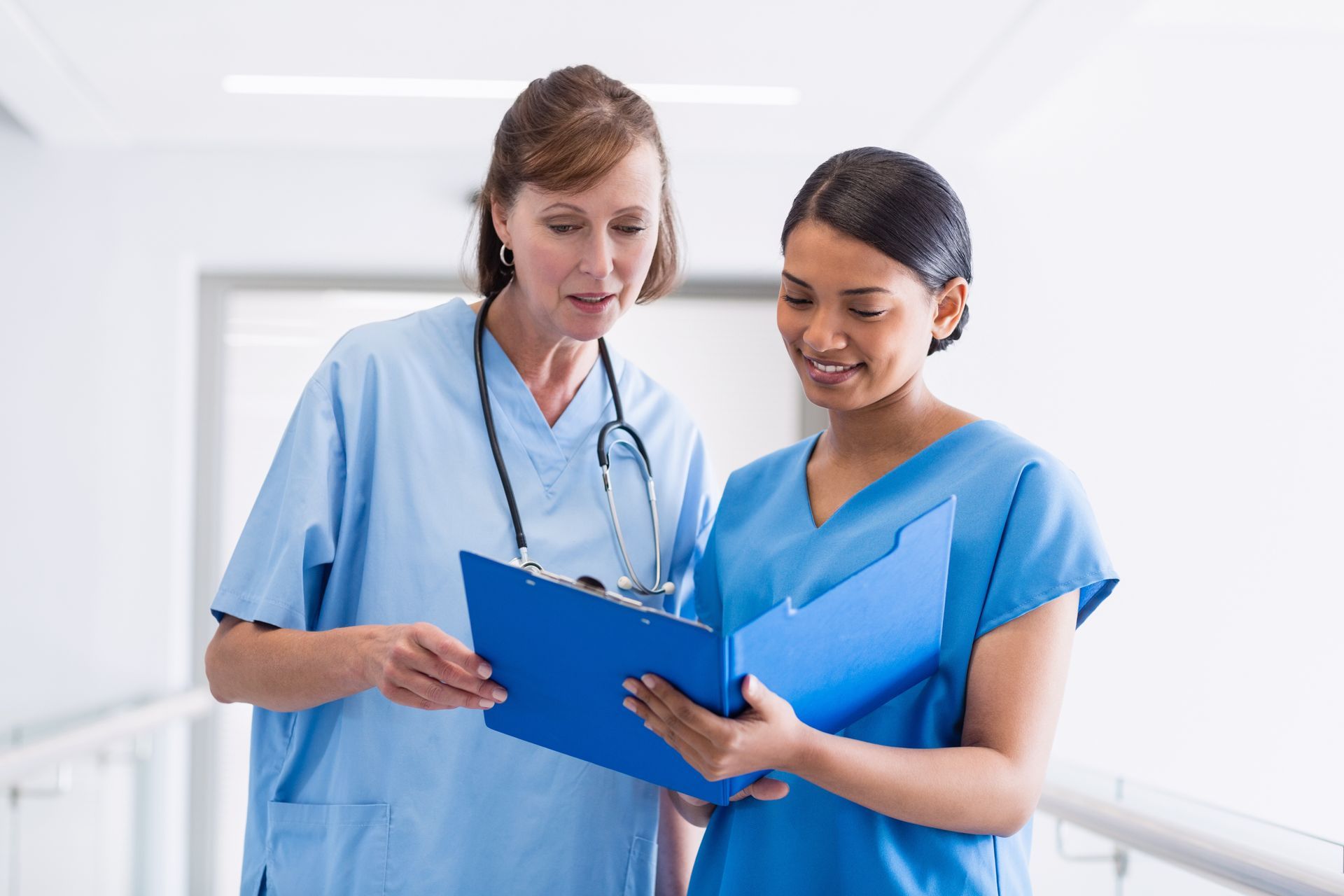 Two nurses are looking at a clipboard in a hospital hallway.