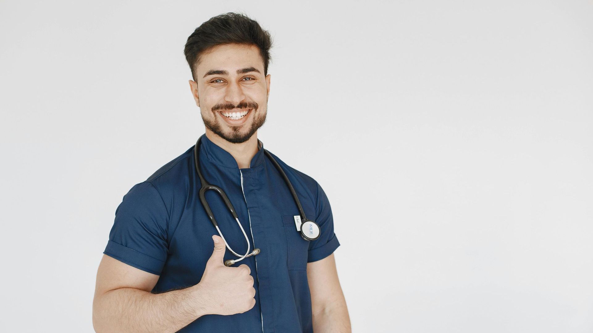 A smiling doctor with a stethoscope around his neck is giving a thumbs up.