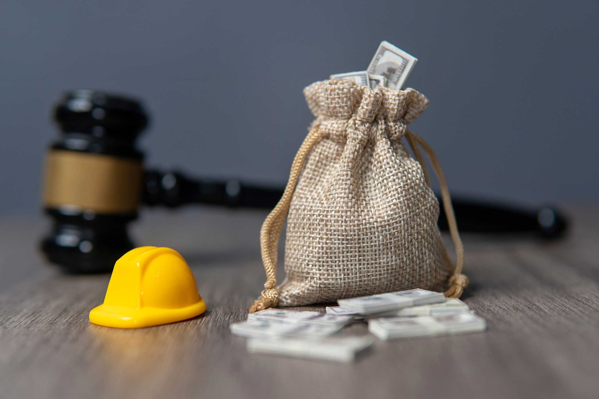A small hard hat and a money bag sit on a table with a gavel blurred in the background. A small hard hat and a money bag sit on a table with a gavel blurred in the background.