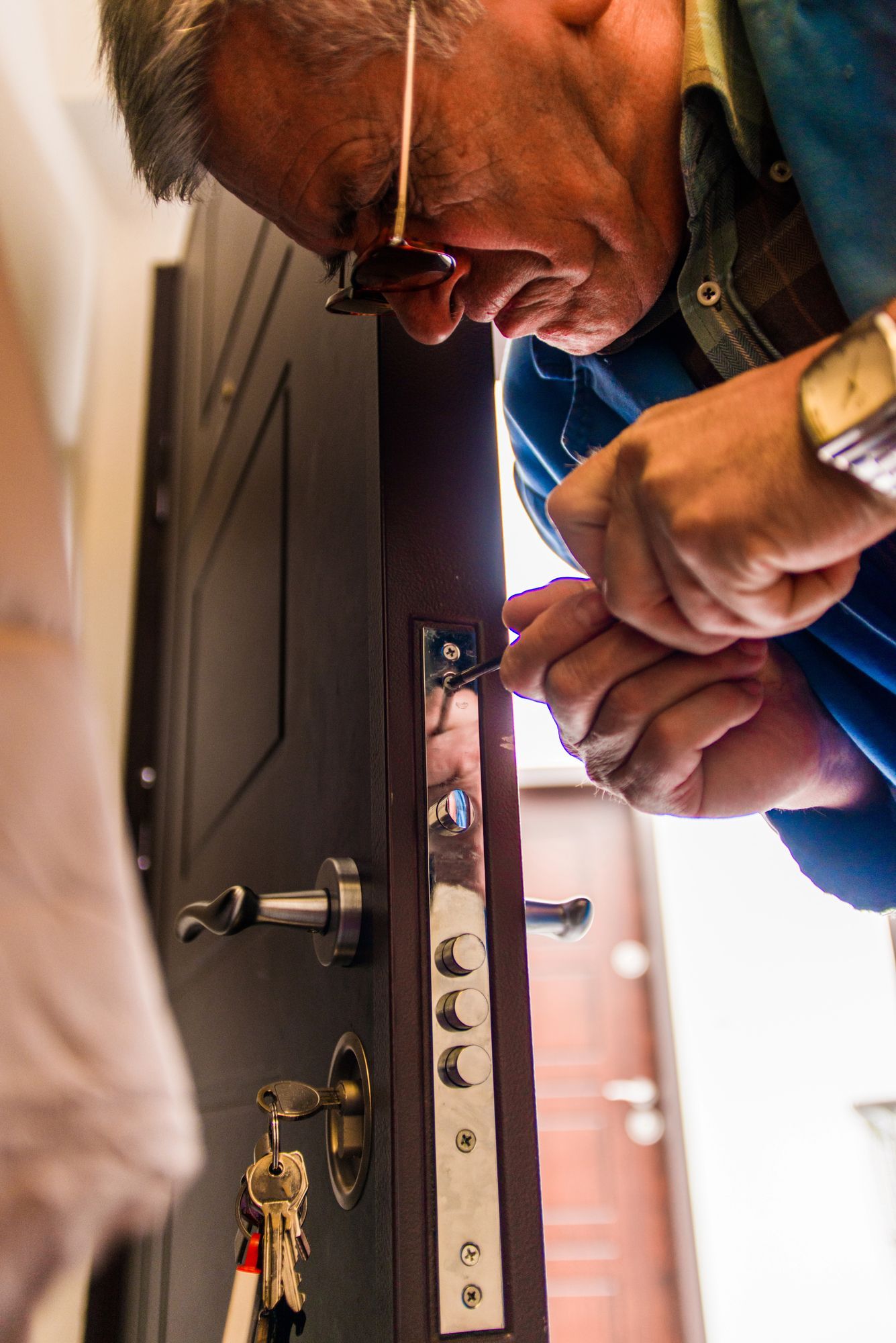 Un hombre con camisa azul usa un destornillador para arreglar la cerradura de una puerta. Una llave cuelga del pomo.