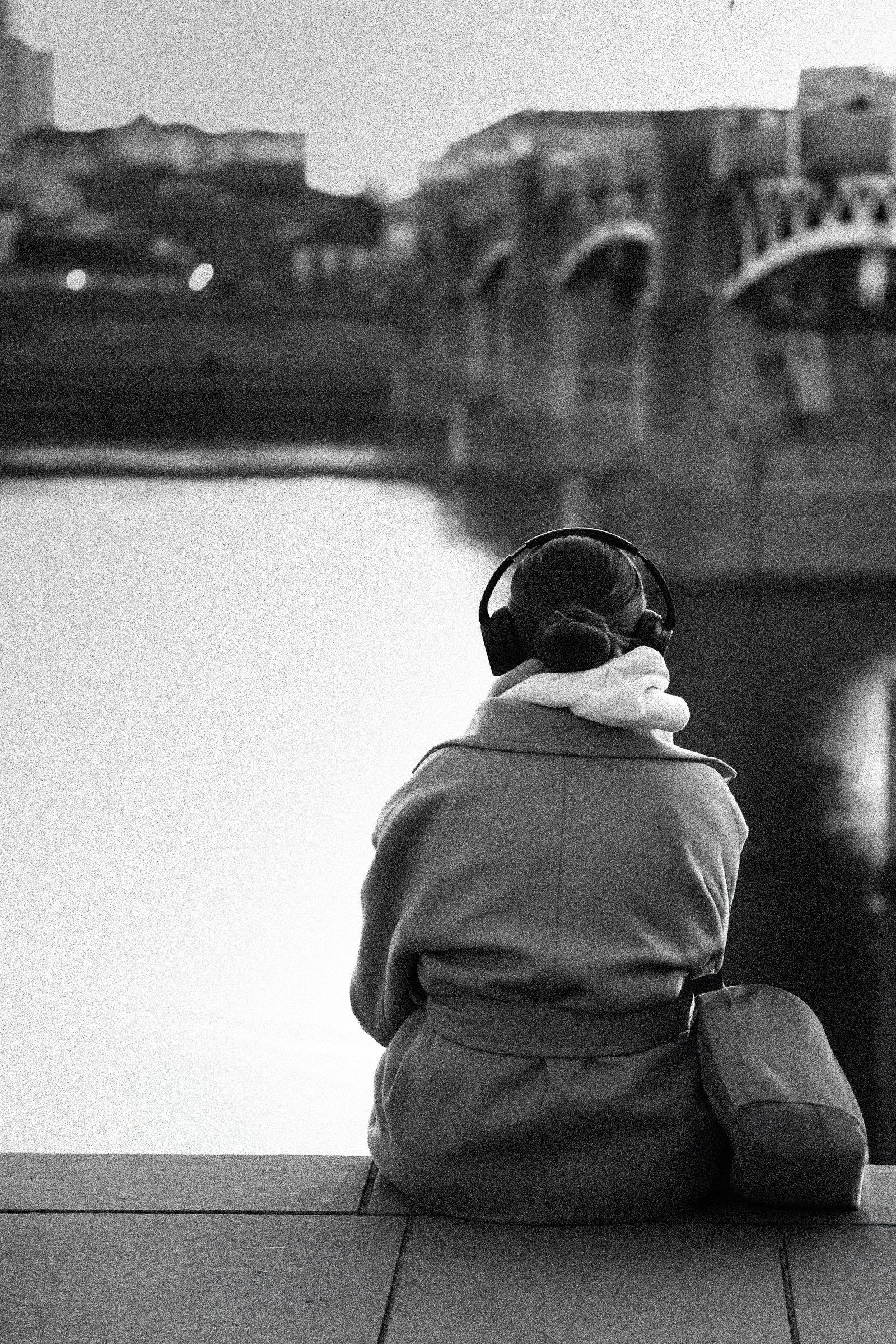 Une personne portant des écouteurs est assise sur un pont surplombant une rivière et des bâtiments en noir et blanc.